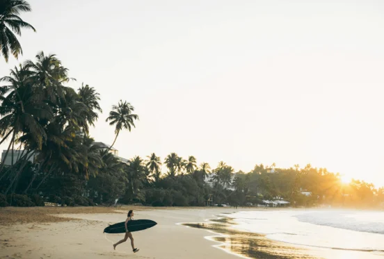 Surfer walking on a palm-lined beach, featured by Ceylon Canvas Holidays.